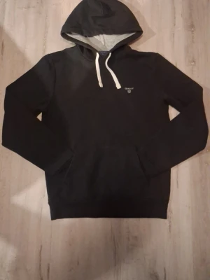 Svart hoodie från GANT med huva - Snygg svart hoodie från GANT med vit dragsko och liten logga på bröstet. Hoodien har en klassisk känguruficka och insidan av huvan är ljusgrå. Perfekt för en avslappnad och stilren look.