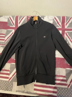 Svart zip tröja från GANT - Snygg svart tröja från GANT med dragkedja framtill och två stora fickor. Liten GANT-logga broderad på bröstet och ribbade muddar vid ärmslut och nederkant. Tillverkad i mjukt bomullsmaterial för en skön känsla. Storlek S 