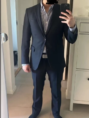 Oscar Jacobson kostym - Klassisk mörkgrå kostymset från Oscar Jacobson. Kavajen har två knappar framtill och fickor med lock. Stilren design med smal passform och diskret struktur i tyget. Perfekt för dig som vill ha en snygg och tidlös look. Både kavajen och byxorna är i utmärkt skick och har inga defekter. Kostymen är bara använd ett fåtal gånger. Skriv gärna för ytterligare frågor. Den passar ca 175 cm.