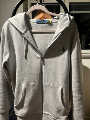 Grå zip hoodie från Polo Ralph Lauren - Snygg grå hoodie från Polo Ralph Lauren med dragkedja, två stora fickor framtill och klassisk huva med dragsko. Ikoniska loggan broderad på bröstet. Tillverkad i mjukt bomullsmaterial som är skönt att ha på sig.