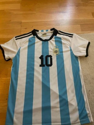 Argentina tröja - Säljer en klassisk Argentina matchtröja från Adidas med ljusblå och vita ränder, svarta detaljer och nummer 10. På bröstet finns AFA-emblemet och på ryggen står det Messi och 10. Perfekt för fotbollsfans! Materialet är lätt och andas.