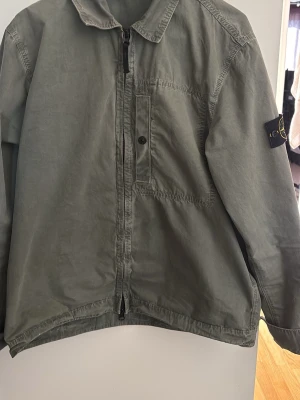 Grön overshirt från Stone Island - Snygg grön overshirt från Stone Island med dragkedja framtill och en bröstficka med knapp. Klassisk krage och ikonisk patch på vänster ärm. Tillverkad i ett slitstarkt bomullsmaterial som ger en avslappnad men stilren look.