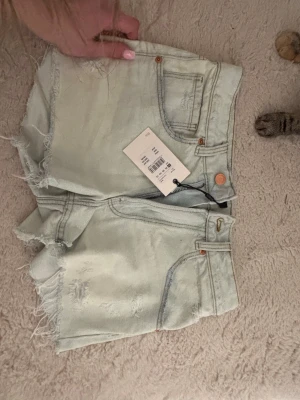 Ljusblå denimshorts med rå kant - Säljer ett par ljusblå denimshorts från H&M med fransig rå kant och slitna detaljer. Klassisk femficksmodell med knapp och dragkedja framtill. Perfekta för sommaren och riktigt snygga till sneakers eller sandaler.