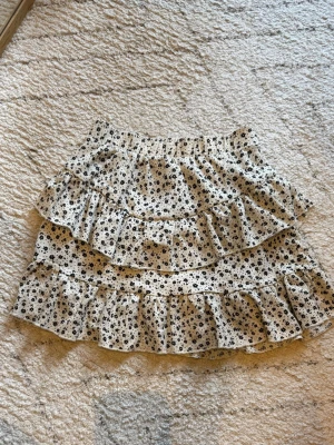 Vit blommig volangkjol - Supersöt kort kjol i vitt med små svarta blommor och flera volanglager. Kjolen har elastisk midja och inbyggda shorts.