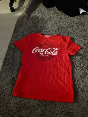 Röd Coca-Cola Classic t-shirt - Säljer en röd t-shirt från Coca-Cola med klassisk logga i vitt framtill. T-shirten har rund halsringning och korta ärmar. Perfekt för dig som gillar retrostil och vill sticka ut med en ikonisk design.