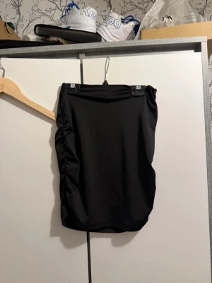 Svart kortkjol med rynkade detaljer - Säljer en svart kortkjol med rynkade detaljer på sidan. Kjolen har en enkel och stilren design som passar till många olika outfits. Materialet känns mjukt och stretchigt för en bekväm passform.