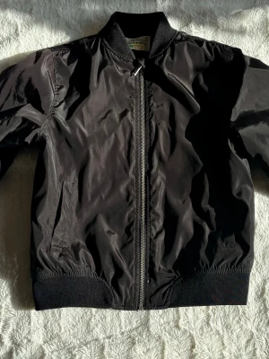 Svart bomberjacka 152 - Stilren svart bomberjacka med klassisk ribbad krage, muddar och nederkant. Jackan har dragkedja framtill och två sidofickor. Tillverkad i glansigt syntetmaterial som ger en cool look.