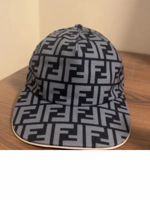 Fendi keps med FF-mönster - Snygg keps från Fendi med ikoniskt FF-logomönster i svart och grått. Klassisk form med böjd skärm och justerbar baksida. Tillverkad i textilmaterial som känns både lyxigt och trendigt. Perfekt accessoar för att lyfta din streetwear-look.