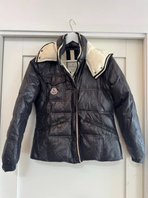 Svart dunjacka från Moncler - Snygg svart dunjacka från Moncler med quiltat mönster, avtagbar huva och vita detaljer på insidan. Jackan har tryckknappar, två snedställda fickor med dragkedja och en bröstficka med Moncler-logga. Storlek 1 motsvarande xs/34. Litet hål på ryggen (sista bilden). Säljes pga för liten.