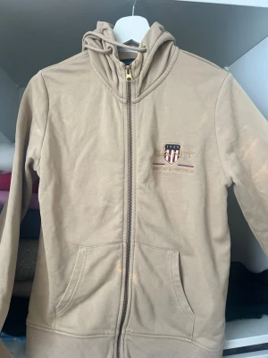 Beige ziphoodie från GANT - Beige hoodie med dragkedja från GANT. Har huva med dragsko, två fickor framtill och broderad logga på bröstet. Perfekt för en avslappnad och stilren look. Mjuk bomullskänsla och ribbade muddar.