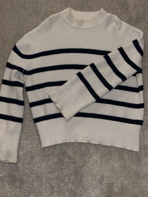 Randig stickad tröja från NA-KD - Säljer en krämvit stickad tröja från NA-KD med svarta horisontella ränder. Tröjan har en rak passform, ribbstickad struktur och rund halsringning. Perfekt för dig som gillar klassisk och enkel stil.
