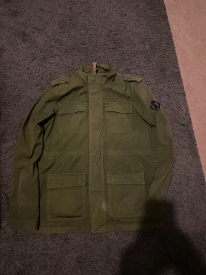 Olivgrön vindjacka med fickor - Snygg olivgrön vindjacka i militärstil med fyra stora fickor framtill, axelklaffar och ståkrage. Jackan har en patch på ena ärmen och stängs med dragkedja och knappslå. Tillverkad i slitstarkt bomullstyg, perfekt för en streetwear-look.