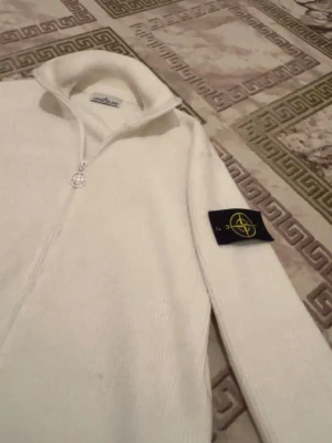 Vit stickad ziptröja från Stone Island - Säljer en stilren vit stickad ziptröja från Stone Island med hög krage och dragkedja framtill. Tröjan har den klassiska Stone Island-loggan på ärmen och är perfekt för lager-på-lager-stil.