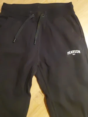 Svarta mjukisbyxor från Pearson - Säljer ett par svarta mjukisbyxor från Pearson med dragsko i midjan och fickor på sidorna. Byxorna har tryckt logga på ena benet och är tillverkade i mjukt material som passar perfekt för chill dagar. Klassisk passform och stilren look.