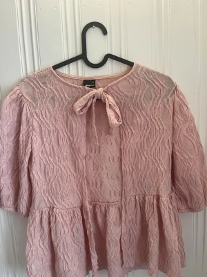 Rosa spetsblus med rosett från Gina Tricot - Söt ljusrosa blus från Gina Tricot i spetsmaterial med strukturmönster. Blusen har trekvartsärm, rund halsringning och en dekorativ rosett framtill (jag har tagit bort 2 rosetter) Den har en lätt volang nedtill och är perfekt till festlig outfit.