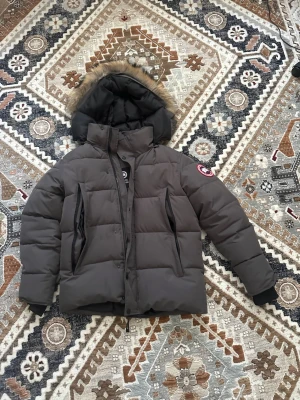 Mörkgrå dunjacka från Canada Goose - Säljer en mörkgrå dunjacka från Canada Goose med klassisk patch på ärmen. Jackan har stor huva med fuskpäls, två snedställda dragkedjefickor framtill och quiltad design. Perfekt för kalla vinterdagar och riktigt varm. Använd bara 2-3 gånger, kan sänka pris bara att skicka ett dm 