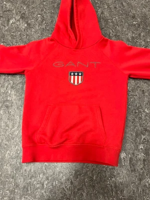 Röd hoodie från GANT med logga - Snygg röd hoodie från GANT med broderad logga och sköld med stjärnor och ränder på bröstet. Klassisk känguruficka framtill och stor huva. Perfekt för en avslappnad och sportig stil. Mjuk bomullsblandning som känns skön mot huden. Pris kan diskuteras