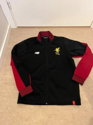 Liverpool träningsjacka New Balance - Snygg svart träningsjacka från New Balance med röda ärmar och krage. Jackan har dragkedja framtill, gul Liverpool FC-logga med texten '125 Years' på bröstet och vit NB-logga. Perfekt för fotbollsfans och sportiga tillfällen.