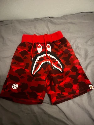 Röda camo shorts från Bape - M men passar S! Säljer ett par röda camo shorts från Bape med den ikoniska hajdesignen framtill. Shortsen har snörning i midjan och är tillverkade i mjuk bomull. Perfekta för dig som vill sticka ut med streetwear-stil.