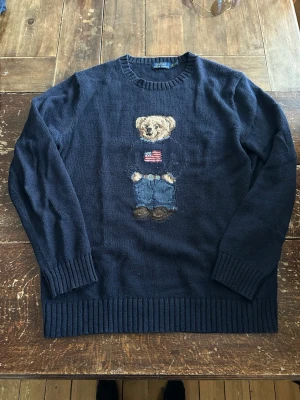 Mörkblå stickad tröja Polo Bear L - Mörkblå stickad tröja från Polo Ralph Lauren med ikoniskt Polo Bear-motiv framtill. Tröjan har rund halsringning, ribbstickade muddar och är tillverkad i mjukt material. Perfekt för dig som gillar klassisk och lekfull stil. size L T. denna är äkta till skillnad på alla bleka Kina fakes som finns på denna app.