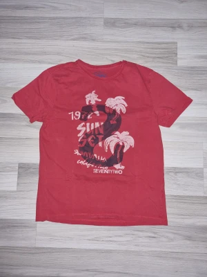 Röd t-shirt med tryck och palmer - Snygg röd t-shirt med coolt tryck framtill som visar palmer och texten 'SUNSET'. T-shirten har rund halsringning och korta ärmar. Perfekt för dig som gillar avslappnad stil och vill sticka ut lite extra.