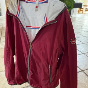 Colmar Jacka  - Snygg vinröd vindjacka från Colmar. Perfekt för dig som vill ha en stilren och lätt jacka. Windbreaker så perfekt till våren och sommaren! Bara att komma med frågor eller funderingar. Den har en liten defekt i luvan men annars bra skick!