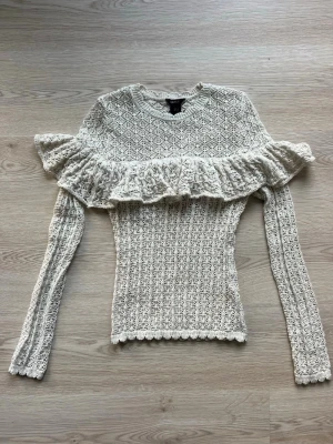 Vit stickad tröja med volang Lindex - Supersöt vit stickad tröja från Lindex med lång ärm och bred volang över bröstet och axlarna. Tröjan har ett detaljerat hålmönster och är figurnära i modellen. Perfekt för dig som gillar romantisk och trendig stil.