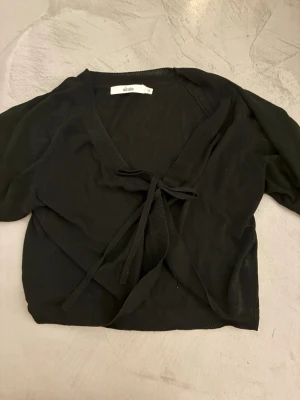 Svart bolero från Aimia - Svart omlottkofta från Aimia med knytband framtill och djup v-ringning.