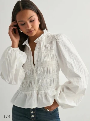 vit topp - Säljer en vit blus med smockdetaljer över bröstet och volang nedtill. Blusen har en klassisk krage, knäppning framtill och långa puffiga ärmar. Materialet ser ut att vara bomull eller liknande och ger en luftig känsla. Perfekt för dig som gillar romantiska detaljer. från nelly nypris 400 