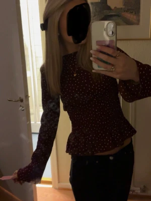 Vinröd prickig blus med volang - Vinröd blus med vita prickar och volang nertill. Långa ärmar och figurnära passform. Perfekt för dig som gillar söta detaljer och ett trendigt mönster.