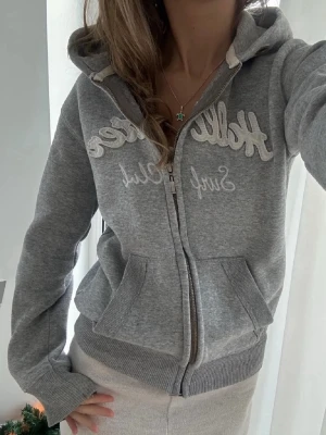 Grå hoodie från Hollister - Snygg grå hoodie från Hollister med dragkedja och broderad logga på bröstet. Mjuk bomullsblandning, två fickor fram och klassisk huva. Perfekt för en avslappnad och trendig stil. Bilden är lånad! 