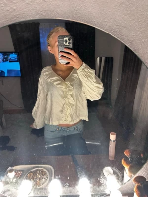 Vit blus med volang och v-ringning - Säljer en vit blus med v-ringning och volangdetaljer framtill. Blusen har långärmade, lätt puffade ärmar och är tillverkad i ett tunt, strukturerat tyg som ger en luftig känsla. Perfekt för dig som gillar en romantisk och trendig stil.