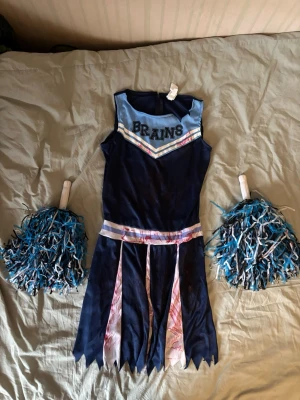 Zombie cheerleader maskeradklänning - Säljer en mörkblå och ljusblå zombie cheerleader-klänning med texten 'BRAINS' på bröstet. Kjolen har blodfläckade detaljer och taggiga kanter. Två blåvita pompoms medföljer.