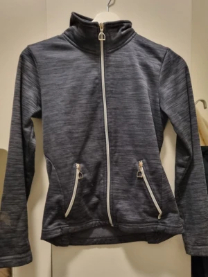 Mörkgrå sportig jacka med dragkedja - Mörkgrå sportjacka i syntetmaterial med hel dragkedja framtill och två snedställda fickor med dragkedjor. Jackan har hög krage och lång ärm, samt diskreta sömdetaljer och metallöljetter bak. Perfekt för träning eller som lager under höst och vinter.