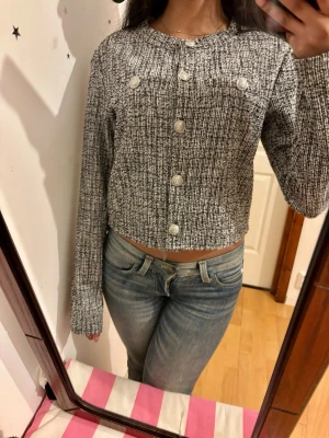 Gråmelerad cropped kofta med knappar - Säljer en gråmelerad cropped kofta med stora vita knappar framtill. Koftan har lång ärm, rund halsringning och ett ribbat stickat mönster. Perfekt för dig som gillar en trendig och avslappnad stil.