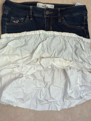 Mörkblå jeansskort från Hollister - Supersnygg jeansskort från Hollister med mörkblå denim upptill och vit, volangig kjoldel i bomull. Klassisk femficksmodell upptill och elastisk midja på kjoldelen. Perfekt för dig som vill ha en mix av jeans och kjol i ett plagg!