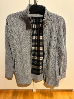 Grå kabelstickad kofta med dragkedja/ fullzip - Snygg grå kabelstickad kofta med dragkedja och hög krage. Insidan har ett rutigt foder i svart, vitt och brunt. Två fickor framtill och ribbade muddar vid ärmslut och nederkant. Perfekt för kyliga dagar och riktigt mysig. Är ingen storlek på lapp men sitter som S. Säljer pga den är för liten. 