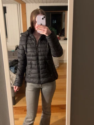 Svart pufferjacka från Navahoo - Svart pufferjacka med huva. Jackan har dragkedja framtill, två fickor med dragkedja och quiltad design. Perfekt för vårdagar och har en sportig look med logotyp på ärmen. Materialet är syntet och jackan är lätt vadderad.