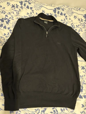 Svart halvzip tröja från BOSS - Snygg svart tröja från BOSS med halv dragkedja och diskret logga på bröstet. Tröjan har ribbade muddar vid ärmslut och nederkant samt hög krage. Perfekt för en stilren och avslappnad look.