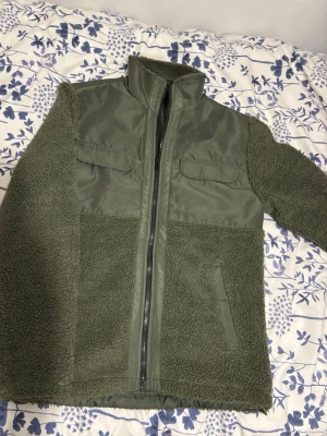 Olivgrön teddyjacka med dragkedja - Snygg olivgrön teddyjacka med dragkedja framtill och hög krage. Jackan har två bröstfickor med lock och två sidofickor. Materialet är en mix av mjuk teddyfleece och slät polyester på bröst och axlar. Perfekt för dig som vill ha en varm och trendig jacka.