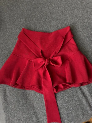 Röd skort med rosett i midjan - Supersnygg röd skort med volang och bred midja, dekorerad med en stor rosett framtill. Kjol framtill och shorts baktill, perfekt för en trendig look. Mjukt tyg som ger en bekväm känsla och snygg passform.
