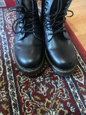 Svarta Dr. Martens stövletter - Klassiska svarta stövletter från Dr. Martens i äkta läder med snörning och den ikoniska gula sömmen runt sulan. Grov, genomskinlig gummisula med bra grepp och hög komfort. Perfekta för dig som vill ha en tidlös och edgy look.