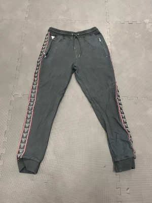 Grå mjukisbyxor från Guess - Snygga grå mjukisbyxor från Guess med svarta och vita logoband längs sidorna och en tunn rosa kant. Byxorna har dragkedjefickor framtill, resår och snörning i midjan samt muddar vid benslut. Perfekta för en chill och avslappnad look.