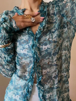  Turkos mesh blus  från Saint Tropez - Snygg transparent blus från Saint Tropez i turkost med blommigt mönster. Blusen har volangdetaljer längs knäppningen och är långärmad med lätt rynkade ärmar. Storlek L men passar mig som är en xs beroende hur man vill den ska sitta. 