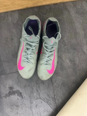 Nike fotbollsskor ljusgrå/rosa - Säljer ett par Nike fotbollsskor i ljusgrått med stora rosa swoosh-loggor på sidorna. Skorna har snörning och en smal, sportig siluett. Ovandelen är tillverkad i ett syntetmaterial som ger bra bollkontroll och passform. Perfekta för dig som vill sticka ut på planen!