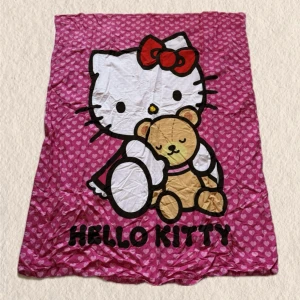 Hello Kitty tyg med nallemotiv - Sött tyg med Hello Kitty och en nalle på framsidan. Bakgrunden är täckt av små rosa hjärtan och texten 'HELLO KITTY' står längst ner. Perfekt för att mysa i soffan eller som dekoration i sovrummet. Skriv för mått ! 1 meter * 1.5meter 