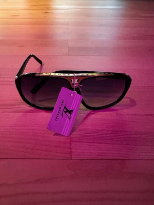 Svarta Louis Vuitton solglasögon - Snygga svarta solglasögon från Louis Vuitton med gulddetaljer och klassisk aviatorform. Bågarna har en elegant guldfärgad detalj med logotyp på sidorna och över näsryggen. Perfekta för att lyfta din stil under soliga dagar.