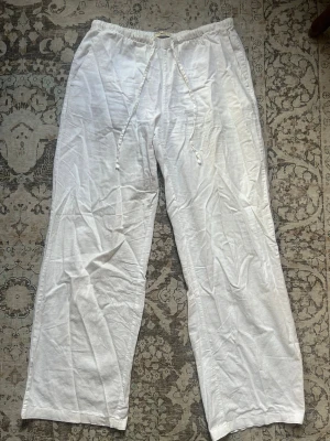 Vita linnebyxor från Gina Tricot - Säljer ett par vita linnebyxor från Gina Tricot med avslappnad passform och dragsko i midjan. Byxorna har raka ben och är tillverkade i ett luftigt linnematerial, perfekta för varma dagar. Klassisk och enkel design som passar till det mesta.