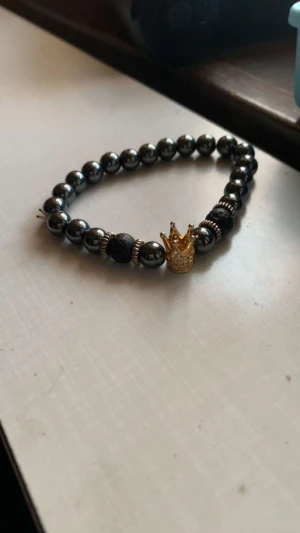 Svart pärlarmband med guldkrona - Stilrent armband med svarta glansiga pärlor och detaljer i silver. Armbandet har en snygg guldfärgad krona som blickfång. Elastisk tråd gör det enkelt att ta på och av. Perfekt accessoar för dig som vill sticka ut med en cool detalj.