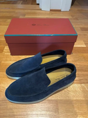 Mörkblå mockaloafers från Loro Piana - Snygga mörkblå loafers i mocka från Loro Piana. Skorna har en klassisk, stilren design med rundad tå och diskret söm längs kanten. Insidan är ljus och mjuk för extra komfort. Perfekta för en clean och avslappnad look. Använda ett fåtal gånger men fortfarande i nyskick. Vid intresse, hör av er för mer bilder eller frågor.